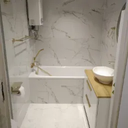 Nowoczesna łazienka z białymi marmurkowymi płytkami, złotą armaturą i drewnianym blatem pod umywalką, widok przez otwarte drzwi.