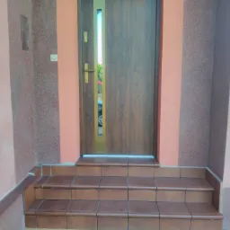 Zewnętrzne schody wejściowe wyłożone brązowymi płytkami ceramicznymi prowadzące do drewnianych drzwi z wąskim przeszkleniem; widoczny fragment elewacji budynku w odcieniach różu i brązu.