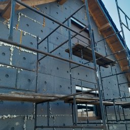 MD-BUD - Dom w trakcie ocieplania styropianem grafitowym, widoczne rusztowanie i pianka uszczelniająca między płytami, szare okna z roletami.