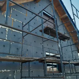 Dom w trakcie ocieplania styropianem grafitowym, widoczne rusztowanie i pianka uszczelniająca między płytami, szare okna z roletami.