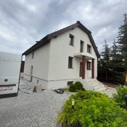 MD-BUD - Dwupiętrowy dom z nową, jasną elewacją, widoczne kolumny wejściowe i schody, obok zaparkowany biały bus, w tle iglaki i pochmurne niebo.