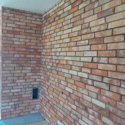 MD-BUD - Ściana wewnętrzna wykończona czerwoną cegłą klinkierową z widoczną instalacją elektryczną i otworem wentylacyjnym, surowy stan pomieszczenia.