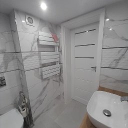 MD-BUD - Wykończona łazienka w bieli i szarości: marmurowe płytki, podwieszana toaleta, grzejnik drabinkowy z ręcznikiem, umywalka nablatowa na drewnianym blacie i białe drzwi z poziomymi frezami.