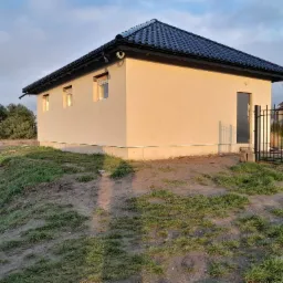Niewielki, parterowy budynek z ciemną dachówką, świeżo otynkowany na beżowo, z trzema małymi oknami i czarnymi rynnami, otoczony nierównym terenem z trawą i ziemią, w tle widoczne drzewa i fragment...