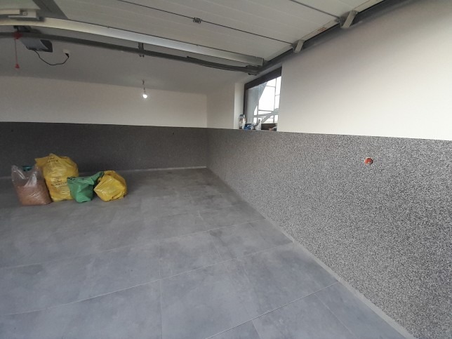 Wnętrze garażu po częściowym wykończeniu: dolna część ścian pokryta szarą, ziarnistą strukturą, górna część biała, widoczne worki z materiałami budowlanymi i szare płytki podłogowe.