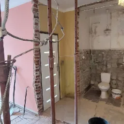 Wnętrze w trakcie prac w Warszawie: podparte metalowymi stemplami otwory w ścianach, widoczne instalacje, resztki gruzu i narzędzia, częściowo usunięte płytki w łazience z toaletą.