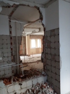 Otwór w ścianie po wyburzeniu, widoczne fragmenty cegieł i gruzu, zarys kolejnych pomieszczeń w tle, naturalne oświetlenie z okna w głębi.