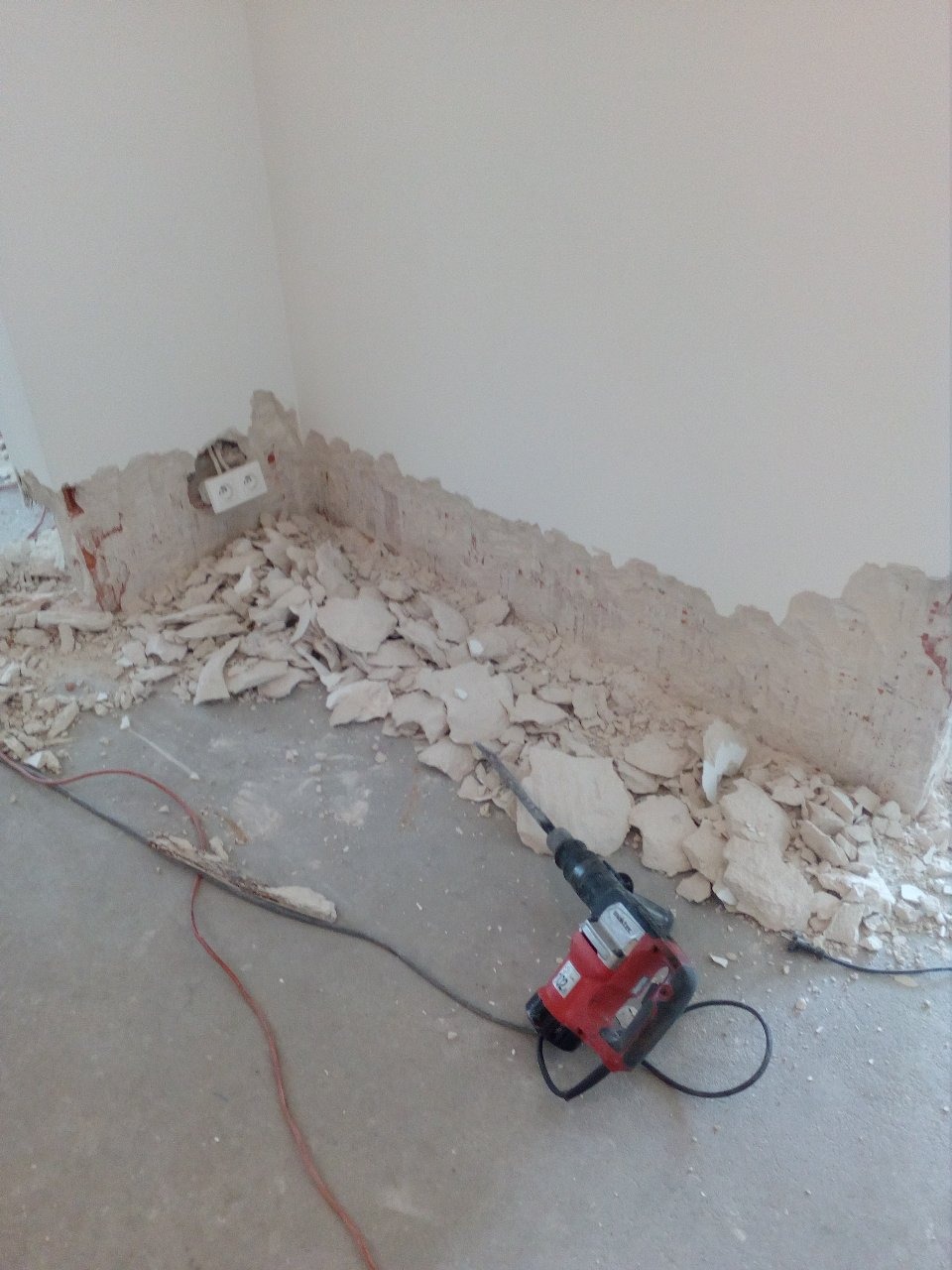 Fragment ściany po wyburzeniu z widoczną puszką elektryczną, gruzem na podłodze i leżącym obok młotem udarowym.