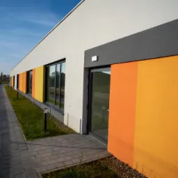 Nowoczesny budynek z pomarańczowo-żółtą elewacją, szarym pasem nad drzwiami i oknami, białą górną częścią ściany oraz szarą kostką brukową na chodniku.