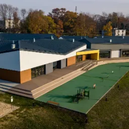 Nowoczesny budynek przedszkola z płaskim dachem, drewnianą elewacją i żółtym wejściem, widok z góry na plac zabaw z piaskownicą i zieloną nawierzchnią.