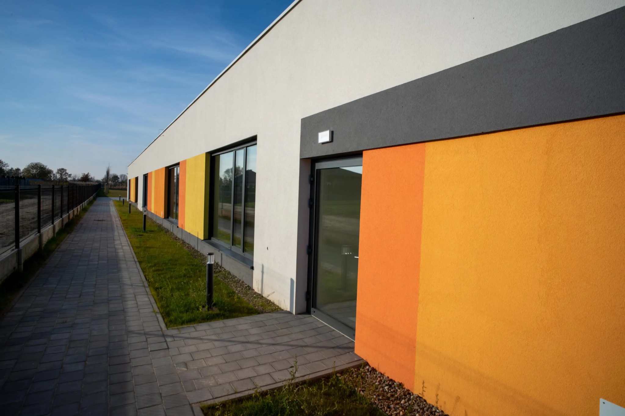 Nowoczesny budynek z pomarańczowo-żółtą elewacją, szarym pasem nad drzwiami i oknami, białą górną częścią ściany oraz szarą kostką brukową na chodniku.