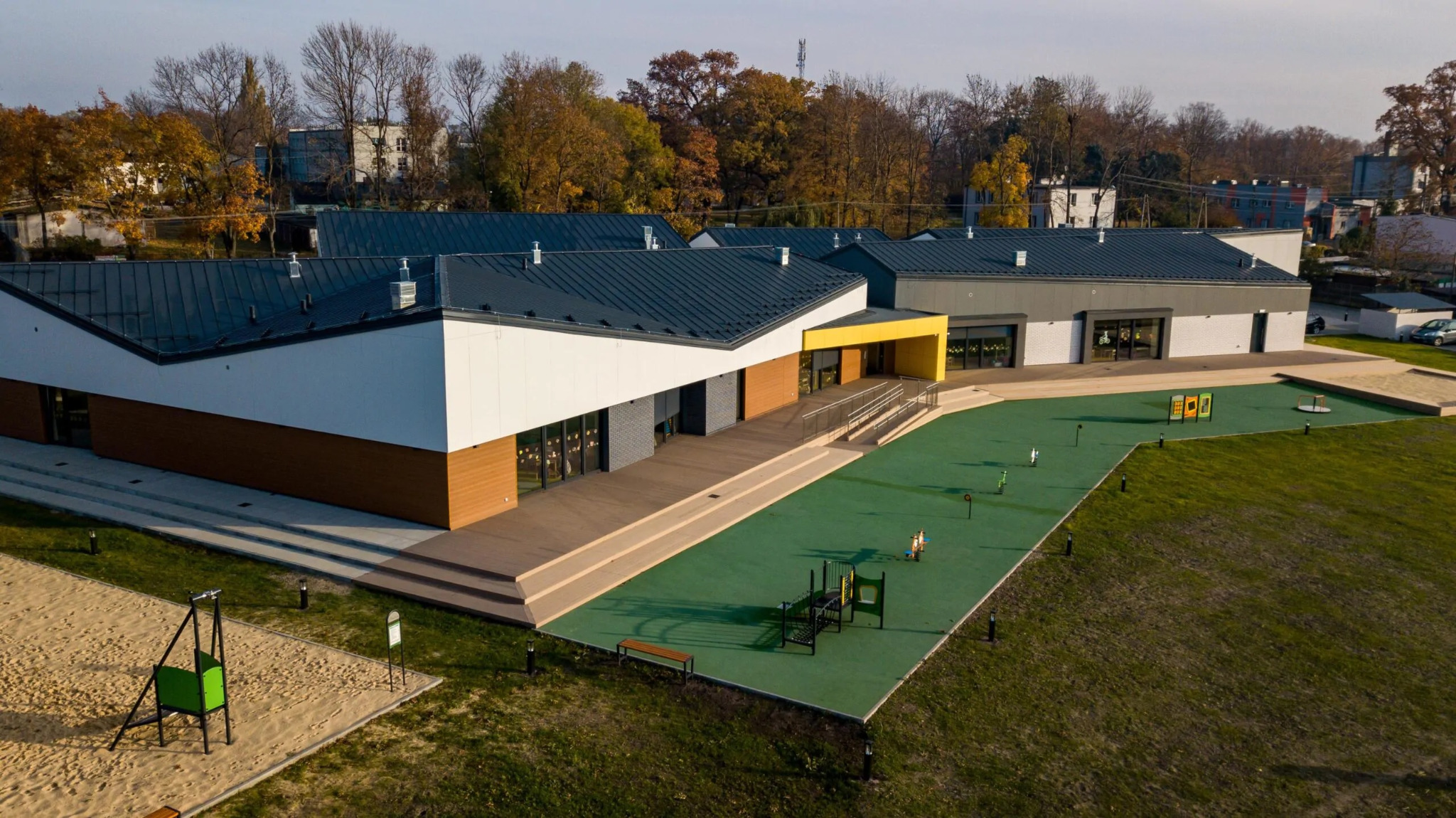 Nowoczesny budynek przedszkola z płaskim dachem, drewnianą elewacją i żółtym wejściem, widok z góry na plac zabaw z piaskownicą i zieloną nawierzchnią.