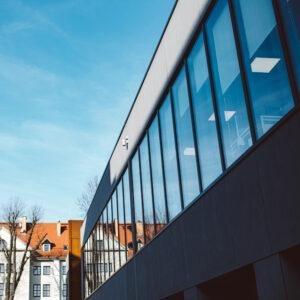 Nowoczesny budynek biurowy z fasadą z długich, prostokątnych okien, widoczny pod kątem od dołu, z fragmentem niebieskiego nieba i budynkami mieszkalnymi w tle.