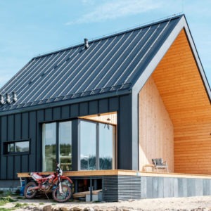 Nowoczesny dom z ciemną elewacją z blachy trapezowej, drewnianym wykończeniem szczytu i przeszklonym narożnikiem, na podwyższonym fundamencie z motocyklem crossowym zaparkowanym obok.