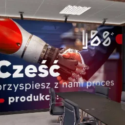 Wnętrze sali konferencyjnej z grafiką na ścianie przedstawiającą uścisk dłoni robota i człowieka, napis 'Cześć, przyspiesz z nami proces produkc'