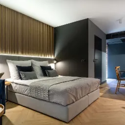 Nowoczesna sypialnia w apartamencie hotelowym z drewnianą ścianą z lameli, szarą tapicerowaną ramą łóżka, niebieską sofą i parkietem ułożonym w jodełkę.