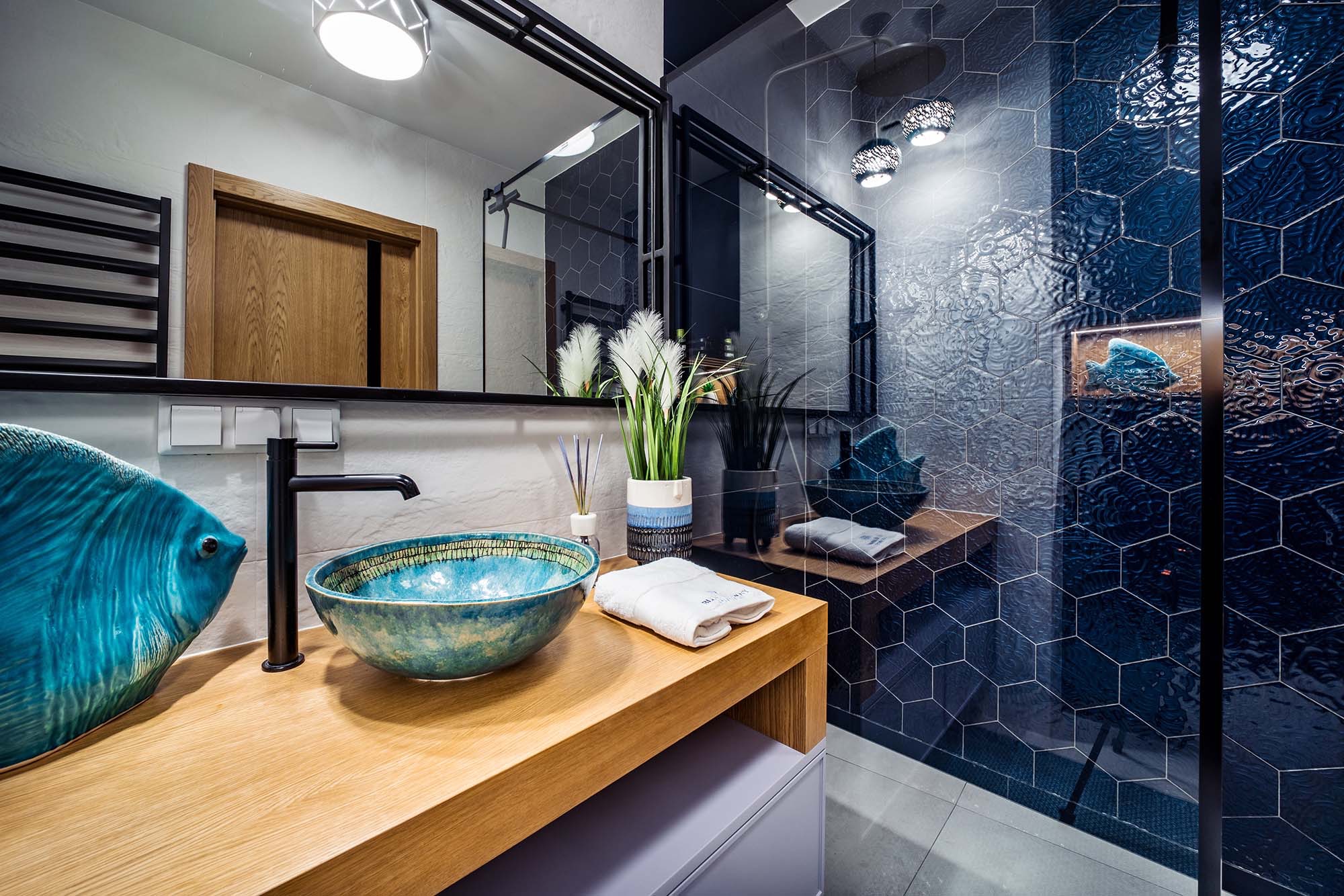 Nowoczesna łazienka w odcieniach błękitu i drewna: ceramiczna umywalka w kształcie misy, dekoracyjna ryba, lustro w czarnej ramie i heksagonalne płytki na ścianie kabiny prysznicowej.