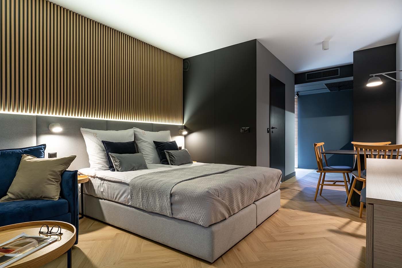 Nowoczesna sypialnia w apartamencie hotelowym z drewnianą ścianą z lameli, szarą tapicerowaną ramą łóżka, niebieską sofą i parkietem ułożonym w jodełkę.