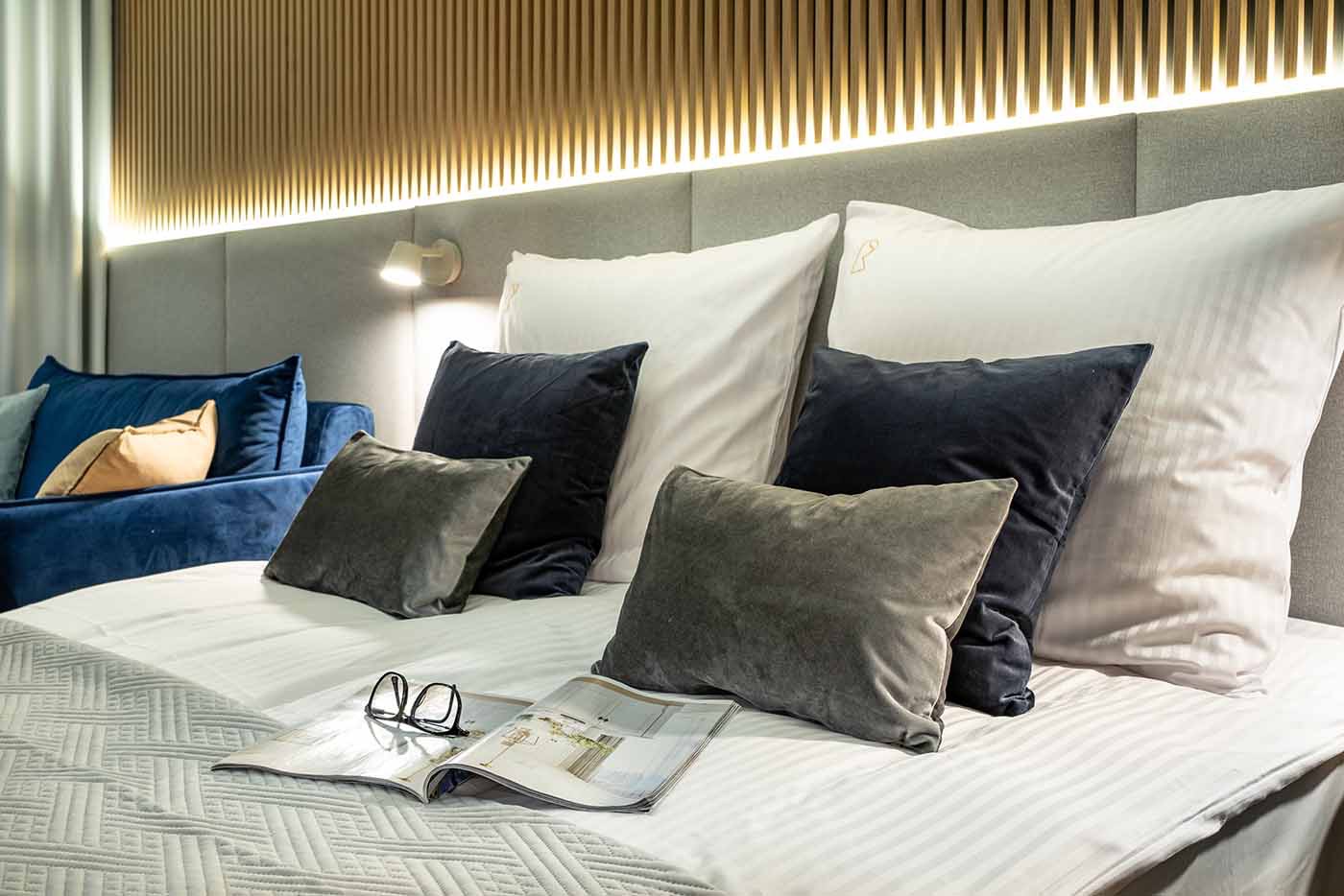 Nowoczesna sypialnia z łóżkiem z szarą tapetą, podświetleniem LED, niebieską sofą w tle, gazetą i okularami na pościeli.