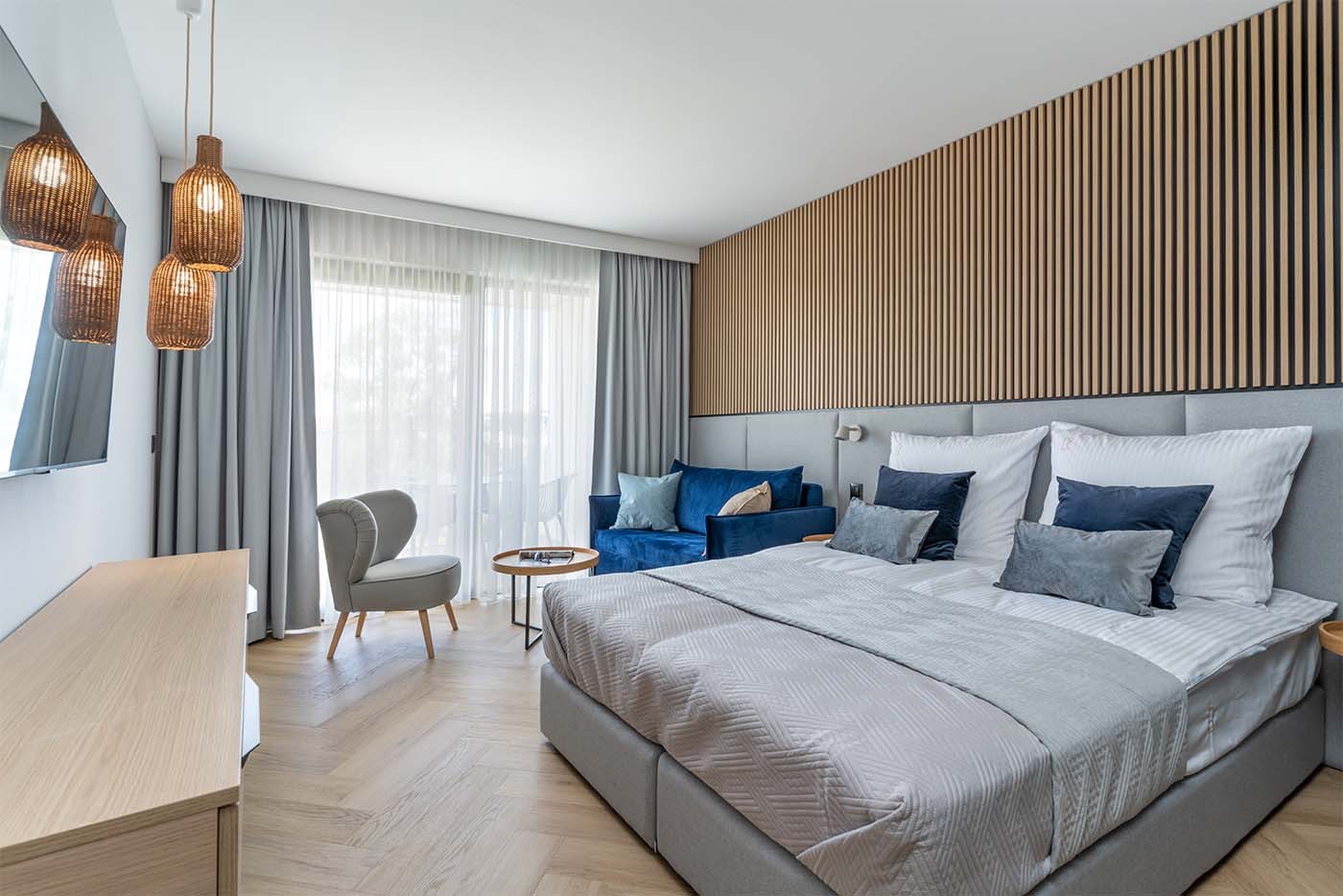 Jasne wnętrze pokoju hotelowego z łóżkiem, sofą i drewnianą ścianą dekoracyjną, oświetlone wiszącymi lampami z plecionki.