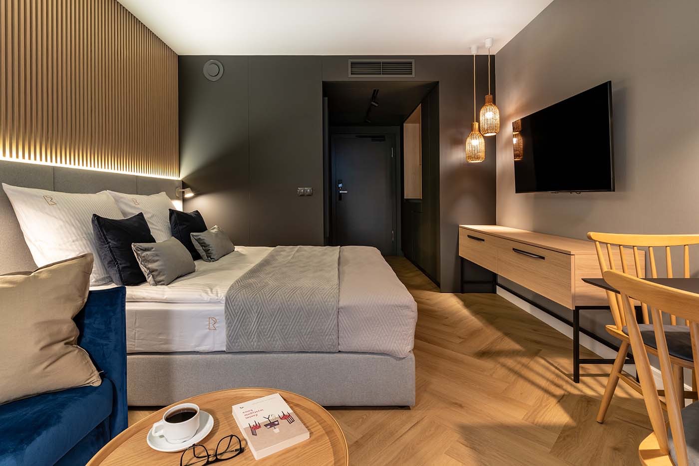 Nowoczesny pokój hotelowy z łóżkiem, drewnianą ścianą z podświetleniem, wiszącymi lampami z abażurami, telewizorem i stolikiem kawowym z książką, okularami i filiżanką kawy.