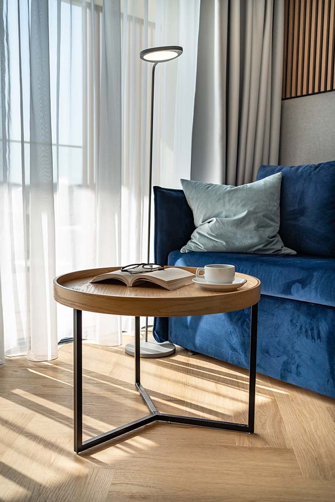 Jasne wnętrze z drewnianym stolikiem kawowym, na którym leży otwarta książka z okularami i biała filiżanka. W tle błękitna sofa i lampa podłogowa.