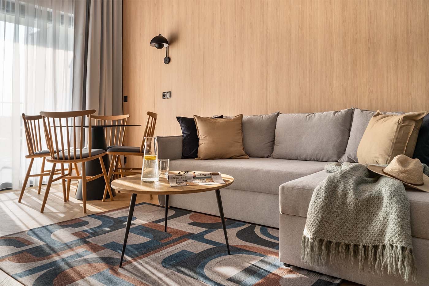 Jasne wnętrze pokoju dziennego z drewnianymi panelami, szarą sofą, okrągłym stolikiem kawowym, czterema krzesłami przy okrągłym stole i wzorzystym dywanem.