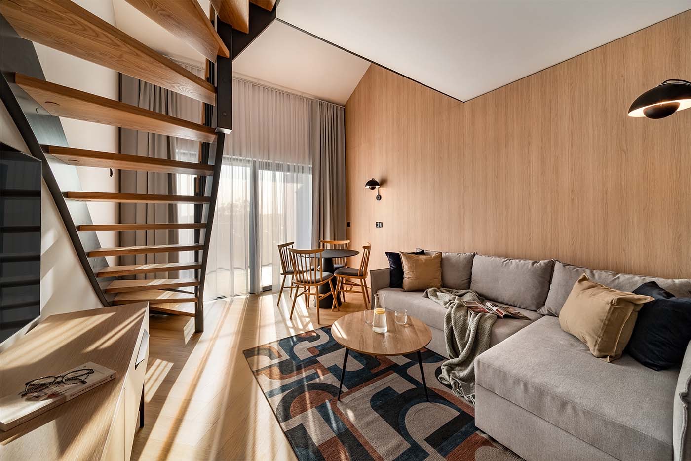 Jasne wnętrze apartamentu z drewnianymi schodami spiralnymi, stołem jadalnym, szarą sofą narożną i wzorzystym dywanem, oświetlone naturalnym światłem.