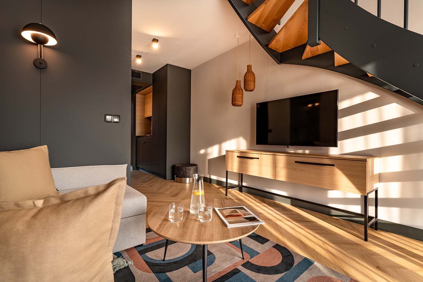Nowoczesny salon z drewnianymi schodami, telewizorem na szafce RTV, okrągłym stolikiem kawowym z wodą i gazetą, oraz designerskimi lampami wiszącymi w kształcie butelek.
