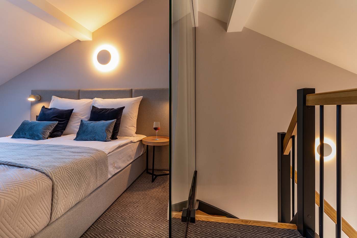 Nowoczesna sypialnia na poddaszu z oświetleniem punktowym, szklaną ścianką działową i schodami z czarnymi balustradami, widok na łóżko z niebieskimi poduszkami i lampką nocną z kieliszkiem wina.