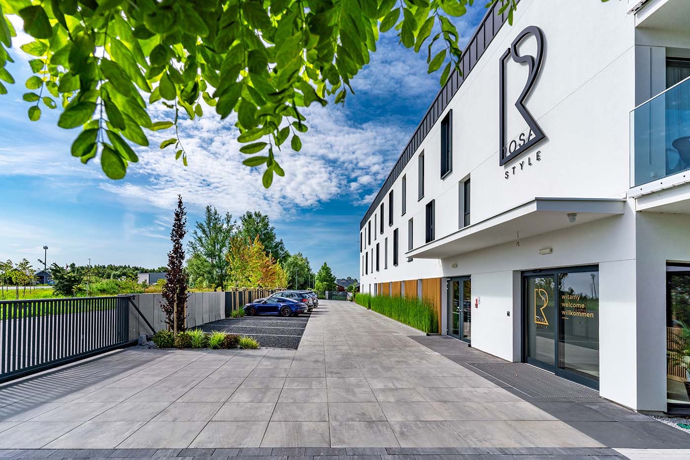 Nowoczesny budynek z logo 'ROSA STYLE' w Trzebiatowie, widoczny z perspektywy parkingu z zaparkowanymi samochodami. Zielone liście drzew w górnej części kadru.