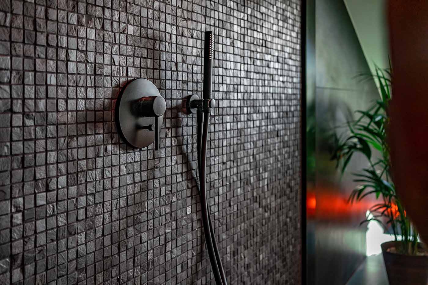 Ciemna mozaika na ścianie kabiny prysznicowej z czarną armaturą i deszczownicą, widoczna roślina doniczkowa w tle.