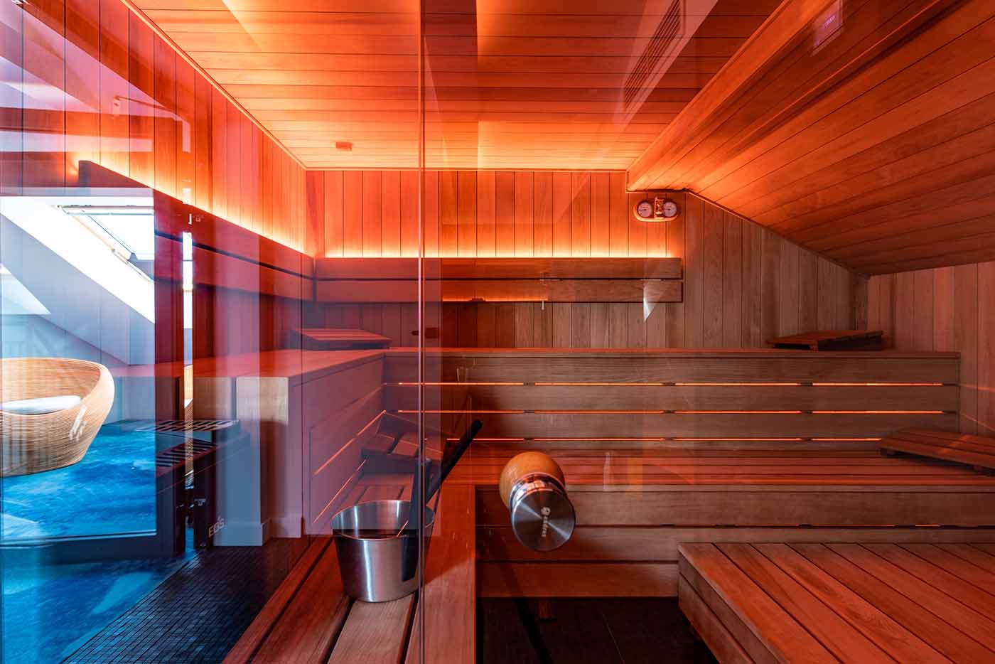 Wnętrze sauny z drewnianymi ławkami, oświetleniem LED i szklaną ścianą, widok na pomieszczenie z wiklinowym fotelem.