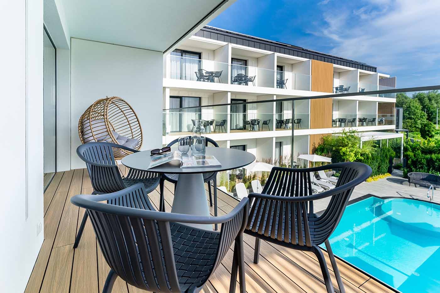 Nowoczesny balkon z drewnianą podłogą, szarymi krzesłami i okrągłym stołem, na którym leżą okulary i gazeta, z widokiem na basen i budynek hotelowy.