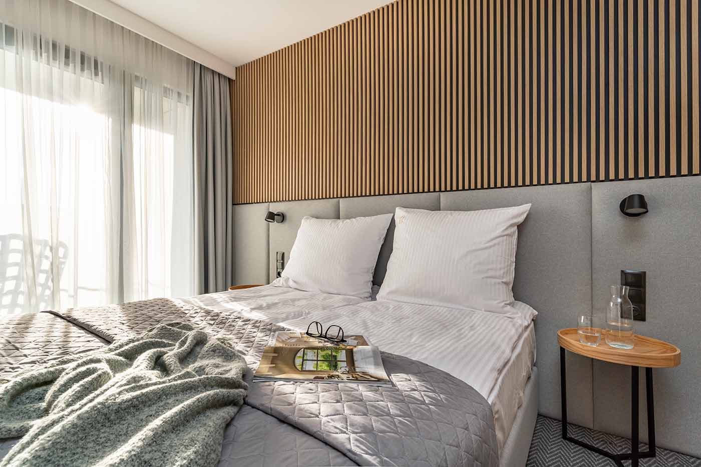 Nowoczesna sypialnia z łóżkiem typu king-size, szarą tapicerowaną ścianą, drewnianą ścianą z pionowych listew, stolikiem nocnym z wodą i szklanką oraz gazetą z okularami na kołdrze.