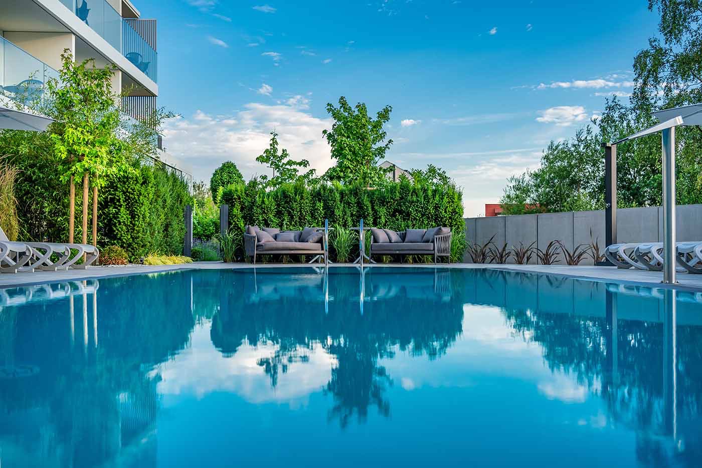 Błękitny basen z odbijającym się niebem, otoczony zielenią i nowoczesnymi meblami ogrodowymi, w tle budynek z balkonami.