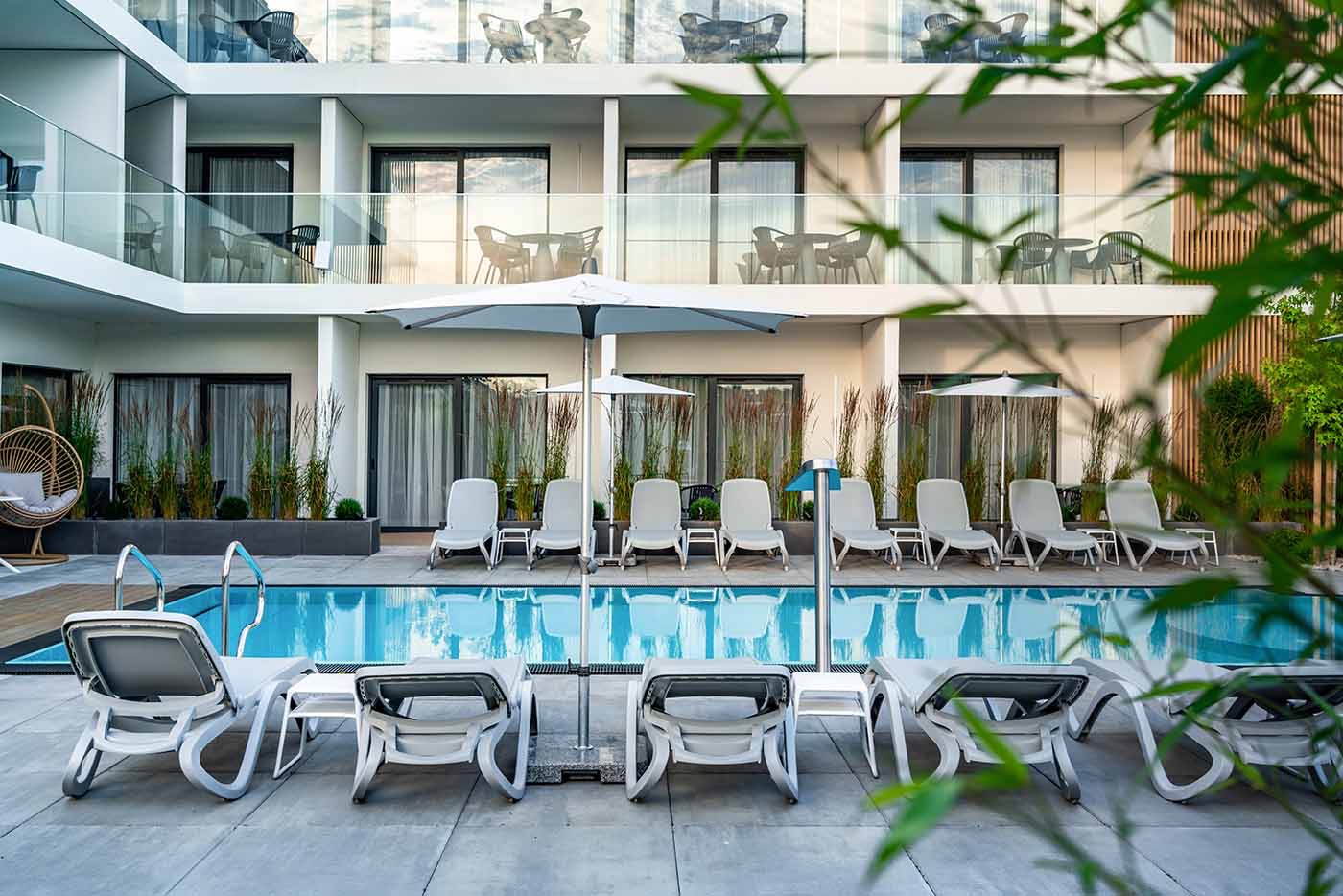 Nowoczesny hotel z basenem i leżakami, widok z zewnątrz, minimalistyczny design, biała fasada, balkony z szklanymi balustradami, parasole przeciwsłoneczne.