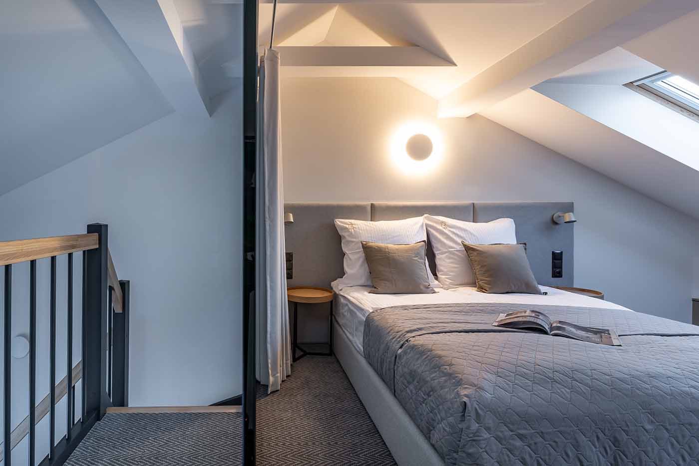 Pokój na poddaszu z łóżkiem, lampą w kształcie okręgu i oknem dachowym, widok z antresoli z czarną balustradą.