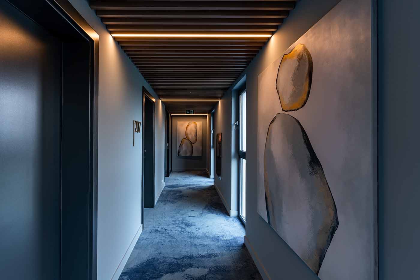 Długi korytarz z szarymi ścianami, niebieską wykładziną i abstrakcyjnymi obrazami na ścianach, oświetlony liniowymi lampami LED w suficie.