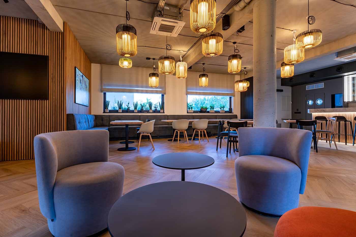 Jasne, przestronne wnętrze kawiarni lub biura z drewnianymi panelami, szarymi fotelami i wiszącymi lampami w kształcie klatek, widok na okno z roślinami w tle.