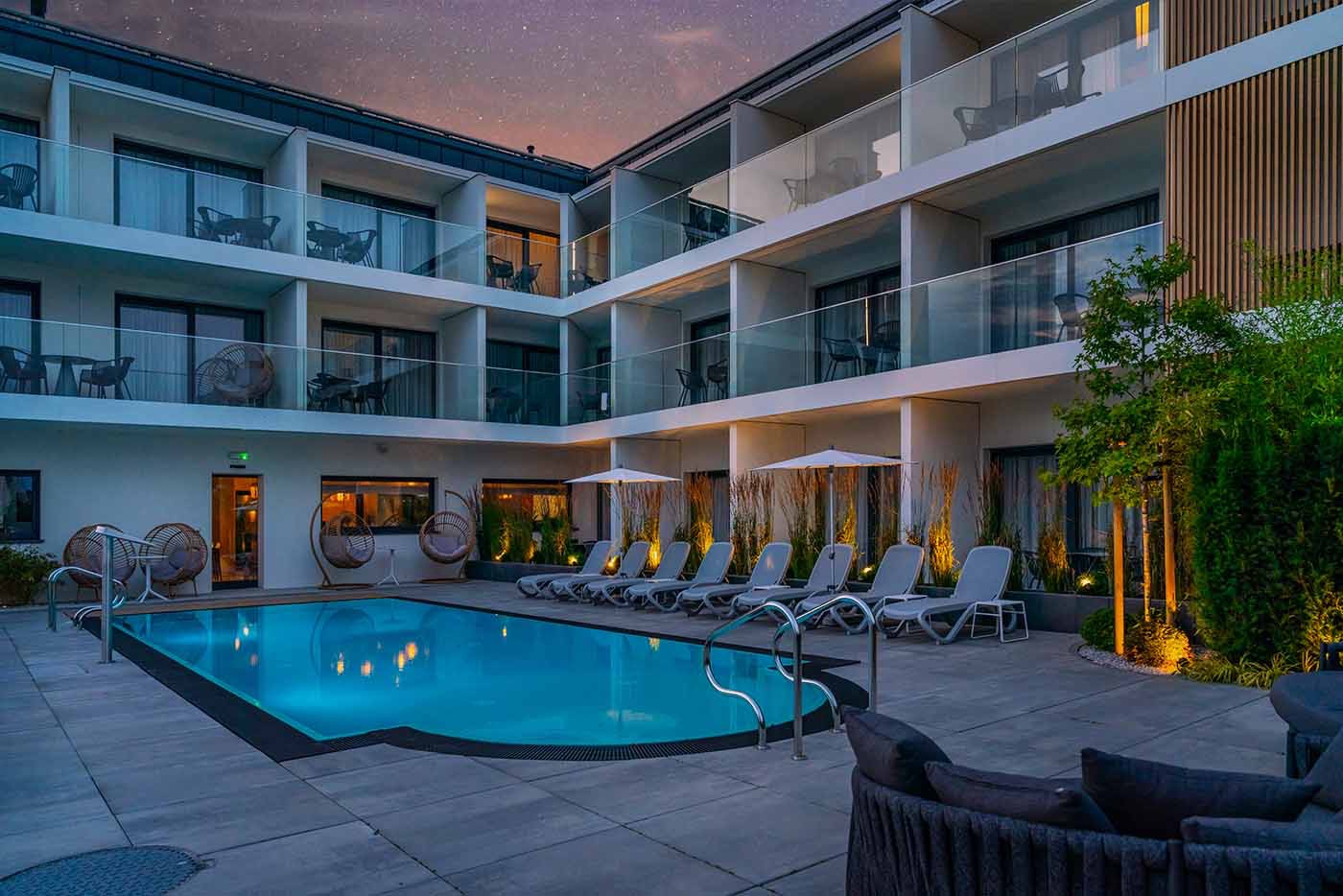 Nowoczesny hotel z basenem oświetlonym nocą, widoczne balkony z krzesłami i stolikami, leżaki przy basenie i starannie zagospodarowana zieleń.