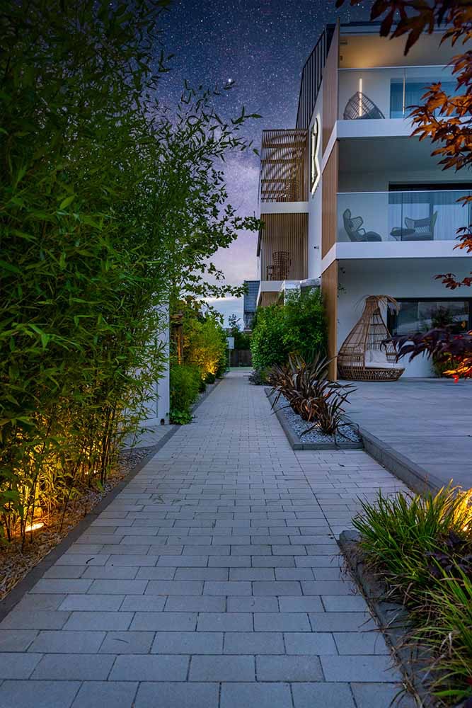 Nowoczesny budynek wielorodzinny z balkonami i oświetloną ścieżką z kostki brukowej prowadzącą wzdłuż bambusowego żywopłotu, nocne niebo w tle.