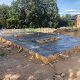 Świeżo wylana, gładka płyta fundamentowa z szalunkiem z płyty OSB, widoczne pionowe rury kanalizacyjne i obudowy pod instalacje, otoczona piaszczystym terenem budowy i zielenią.