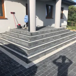Schody wejściowe do domu wyłożone ciemną kostką z jasnymi akcentami, przylegający chodnik z kostki, w tle fragment elewacji budynku z oknami i kolumnami.