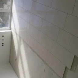 Układanie podłogi z płytek ceramicznych w jasnym odcieniu, widoczne niedokończone fragmenty i ślady zaprawy, okno w tle.