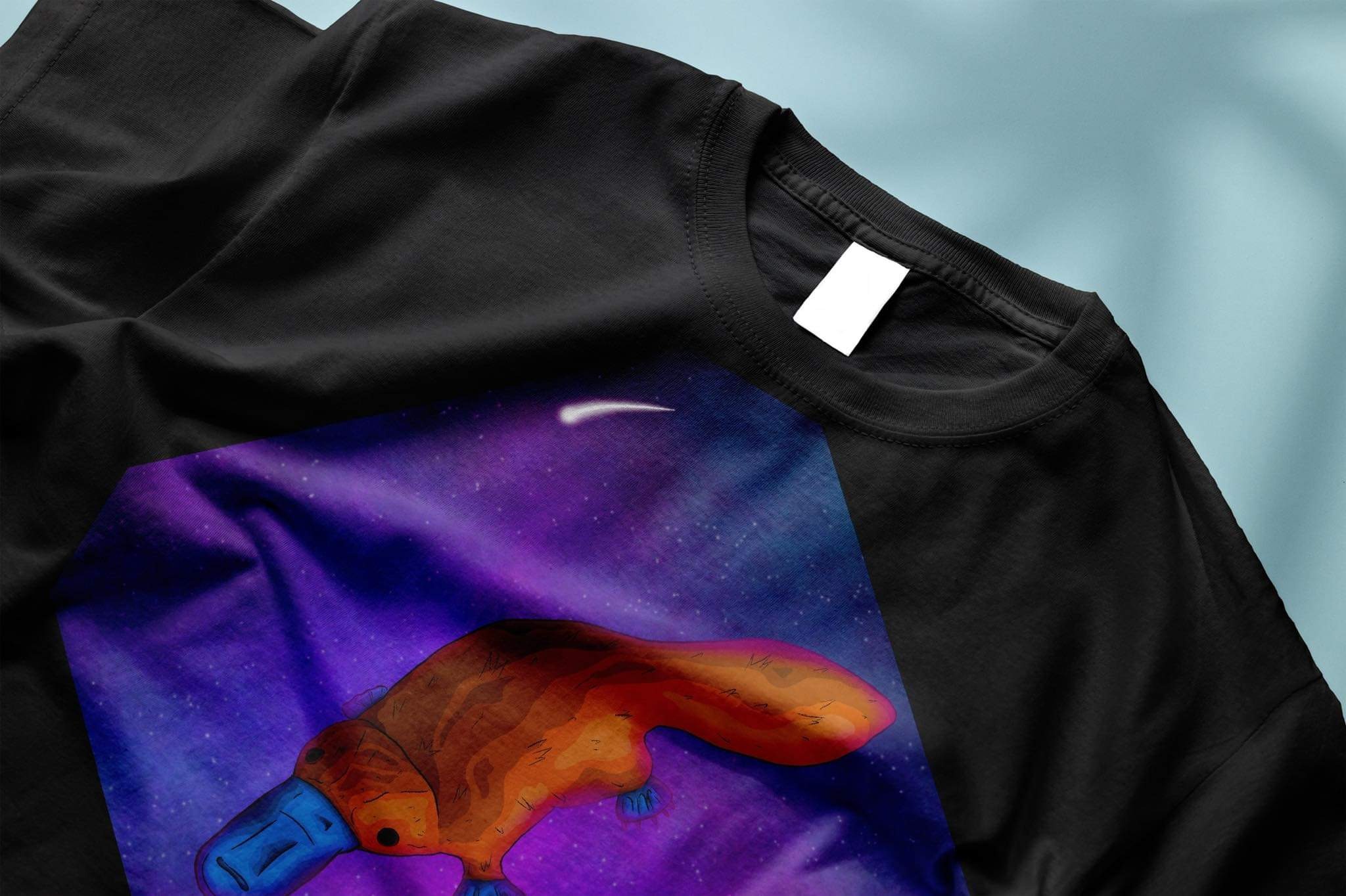 Czarna koszulka z nadrukiem przedstawiającym stylizowanego dziobaka na tle kosmicznego krajobrazu w odcieniach fioletu i granatu, leżąca na jasnoniebieskim tle.