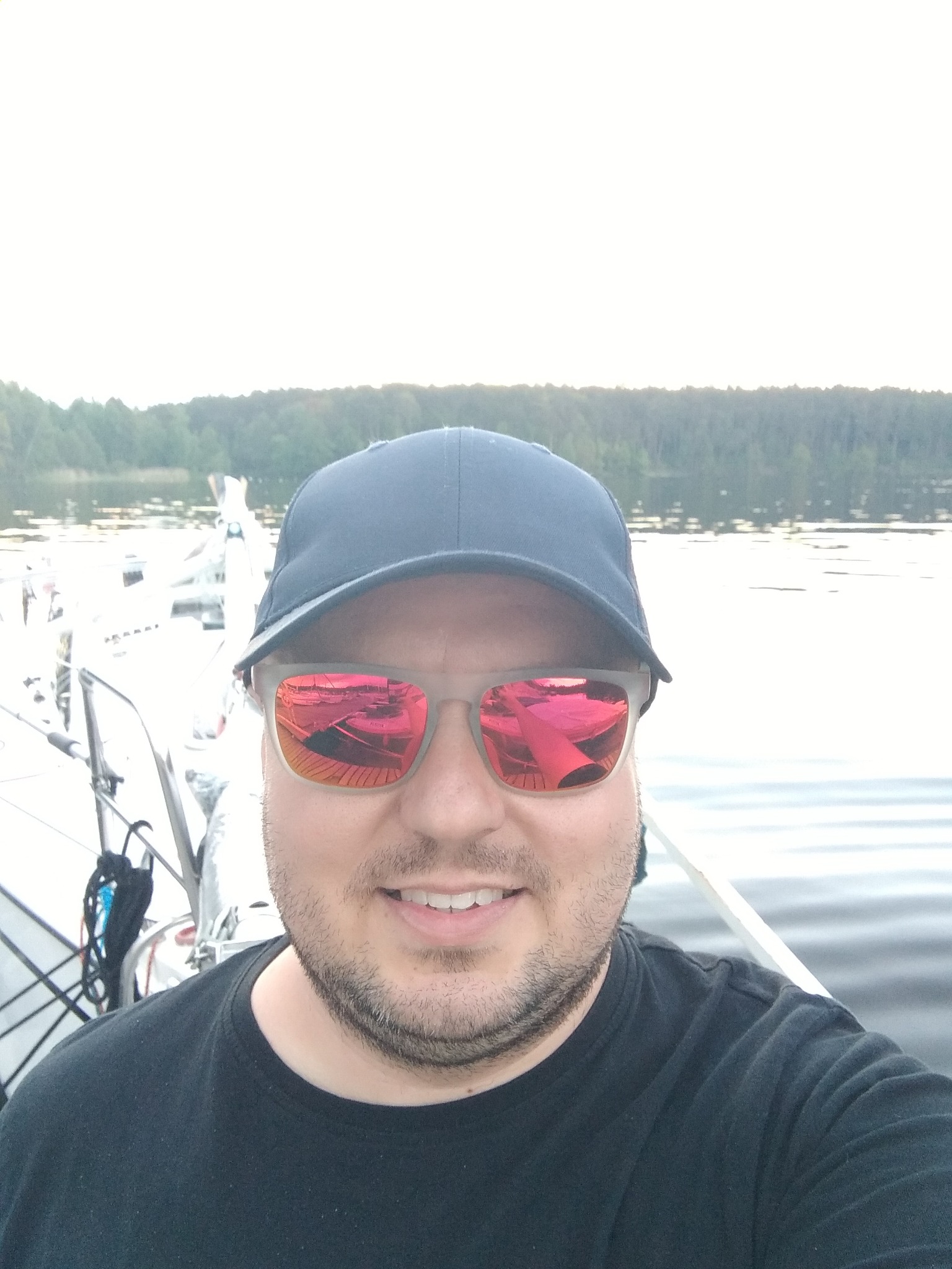Mężczyzna w czarnej koszulce i czapce z daszkiem, w okularach przeciwsłonecznych odbijających łodzie na jeziorze, na tle jeziora i lasu.