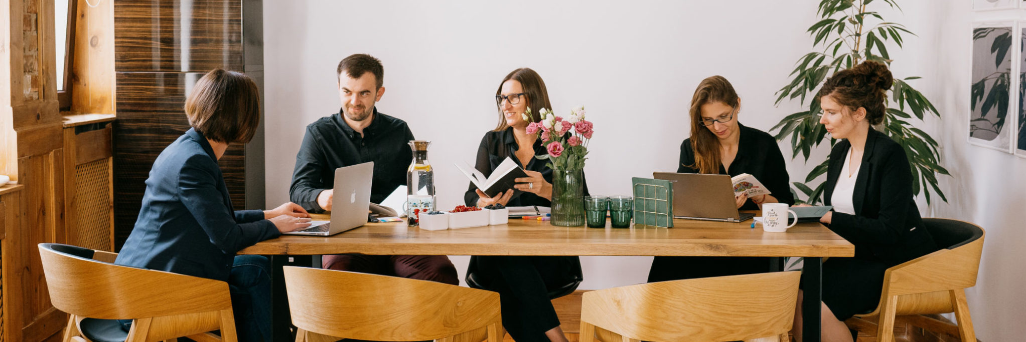 Zespół pięciu osób pracuje przy drewnianym stole, używając laptopów i notatników; na stole wazony z kwiatami, woda z owocami i miseczki z owocami.