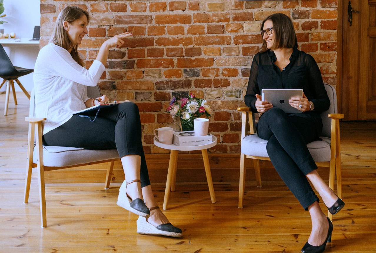 Dwie kobiety siedzą na krzesłach podczas spotkania, jedna z notatnikiem i długopisem, druga z tabletem, na tle ceglanej ściany.