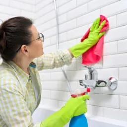 Kobieta w okularach i zielonych rękawicach czyści białe płytki w łazience, używając różowej ścierki i niebieskiej butelki ze spryskiwaczem.